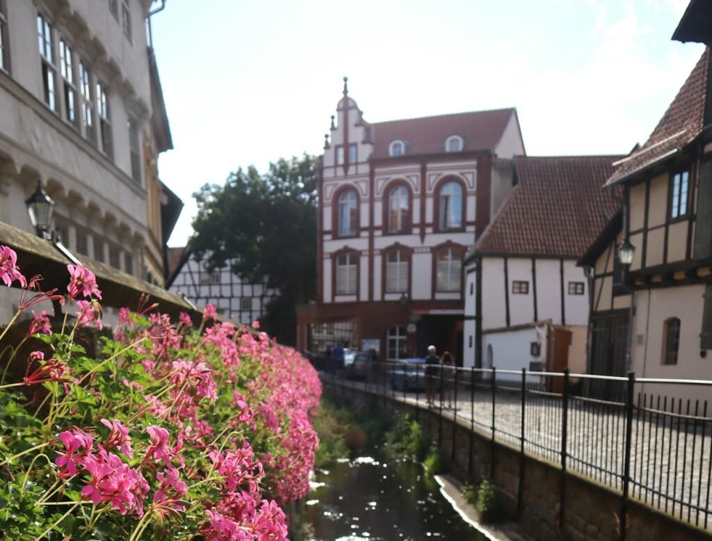 Malerische Aussicht auf ein europäisches Dorf mit einer blumengeschmückten Brücke über einen kleinen Kanal und traditionellen Gebäuden, darunter das Geburtshaus, im Hintergrund.