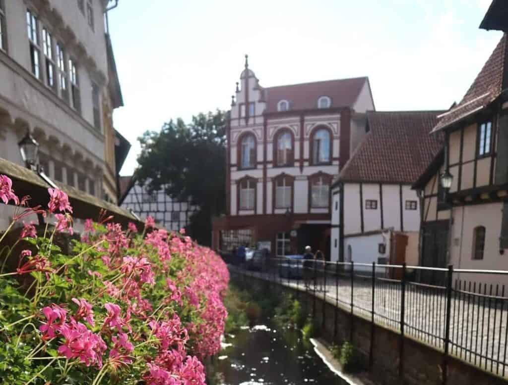 Rosa Blumen säumen einen Metallzaun an einem schmalen Kanal. Im Hintergrund stehen historische Fachwerkhäuser und ein großes weißes Backsteingebäude unter einem teilweise bewölkten Himmel. Die ruhige Szenerie vermittelt Geborgenheit und eignet sich als Symbol für einen geschützten Raum bei Gesprächen rund um Geburtserfahrungen oder Ängste vor einer weiteren Geburt.