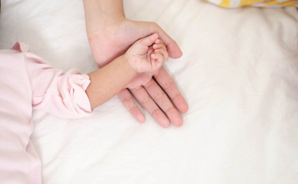Eine erwachsene Hand ruht ruhig auf einer weißen Oberfläche, während eine kleine Babyhand im rosa Ärmel behutsam einen Finger der erwachsenen Person umschließt. Diese Geste vermittelt Nähe und Geborgenheit zwischen Erwachsenem und Kind.