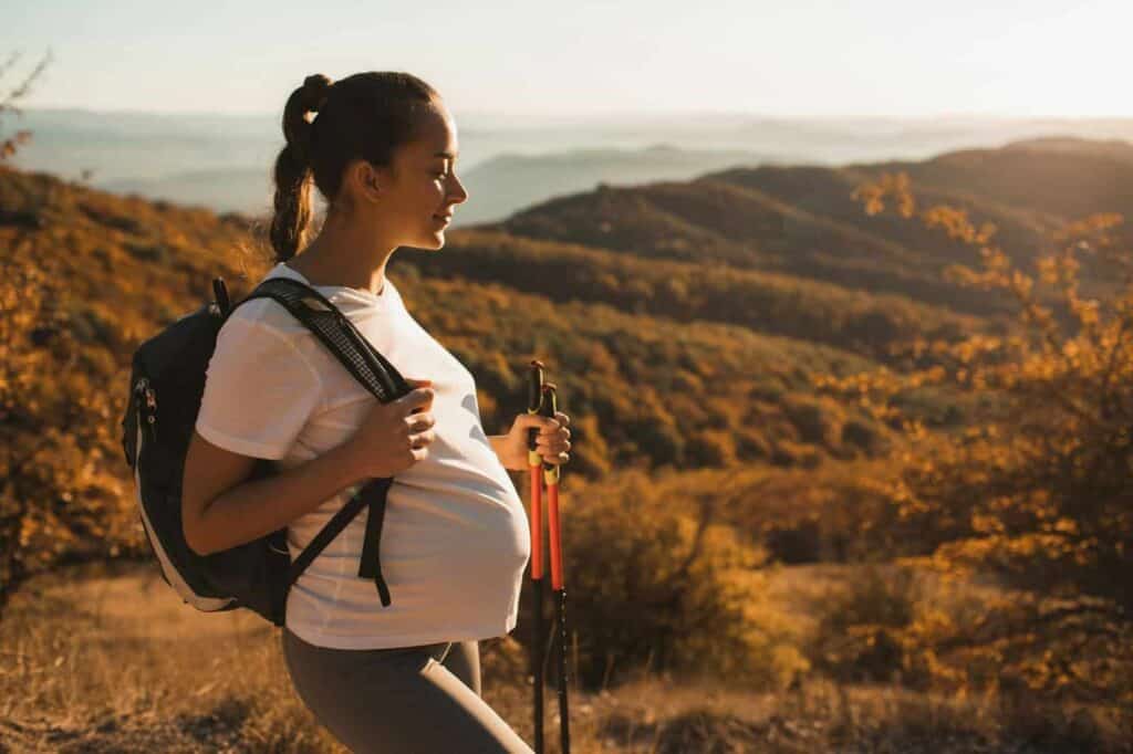 Eine schwangere Frau mit Rucksack und Wanderstöcken steht auf einem Hügel und blickt bei Sonnenuntergang über eine ruhige Landschaft mit Bäumen und fernen Bergen; sie verweilt nachdenklich, geprägt von den Erfahrungen einer Folge-Schwangerschaft nach einer traumatischen Geburt.