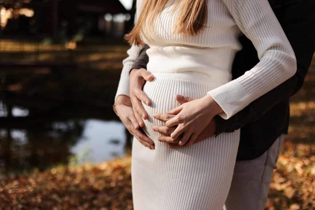Eine schwangere Frau in einem weißen Strickkleid steht draußen. Hinter ihr steht eine weitere Person; beide legen behutsam ihre Hände auf den Babybauch. Diese Geste vermittelt Geborgenheit und einfühlsame Unterstützung während einer Schwangerschaft nach einer schwierigen Geburtserfahrung.