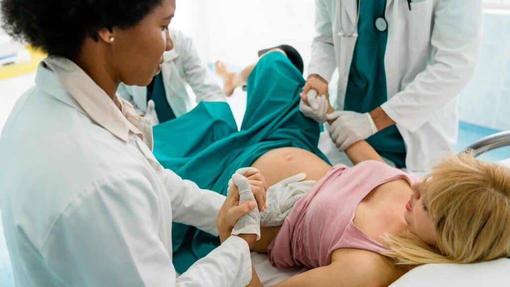 Ein Team aus Ärzt*innen begleitet eine schwangere Frau während der Geburt auf einem Krankenhausbett, unterstützt sie einfühlsam und ist für sie da. Es stehen Informationen zur traumainformierten Beratung zur Verfügung, um insbesondere bei einer weiteren Schwangerschaft nach schwierigen Geburtserfahrungen Unterstützung zu bieten.