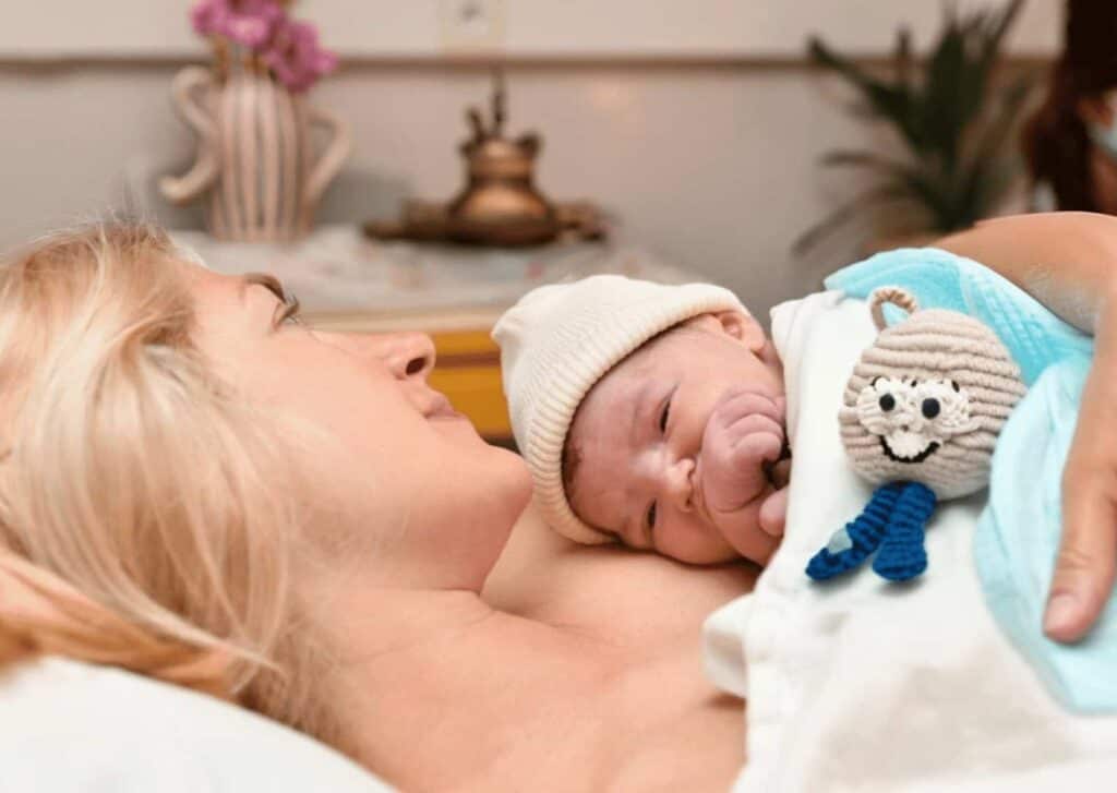Ein Neugeborenes mit einer weißen Mütze liegt, in eine Decke gehüllt, auf der Brust einer Frau. Neben dem Baby liegt ein gehäkeltes Stofftier. Die Szene zeigt eine ruhige, geborgene Situation nach der Geburt, die insbesondere für Menschen nach belastenden Geburtserfahrungen von Bedeutung sein kann.