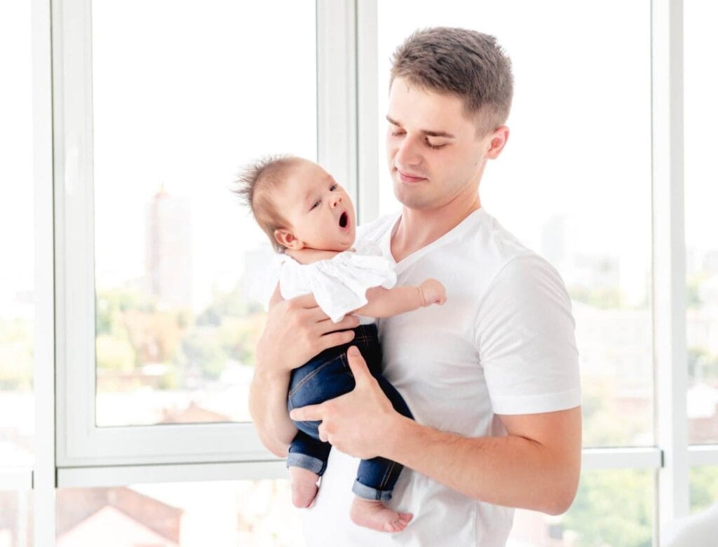 Ein Mann in einem weißen Hemd hält ein gähnendes Baby in einem weißen Oberteil und Jeans auf dem Arm. Beide stehen vor großen Fenstern mit Blick auf eine Stadtlandschaft. Die Szene wirkt ruhig, geborgen und vermittelt die Nähe zwischen Vater und Kind.