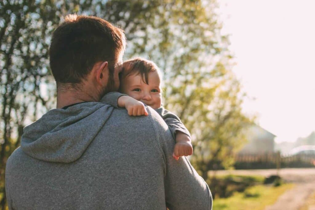 Ein Mann in einem grauen Kapuzenpullover hält ein Baby behutsam auf seiner Schulter, draußen zwischen Bäumen und im Sonnenlicht. Die ruhige Szene strahlt Geborgenheit aus und verweist zugleich darauf, dass nach der Geburt Sorgen oder Ängste auftreten können, etwa vor einer weiteren Geburt oder im Umgang mit belastenden Erfahrungen.