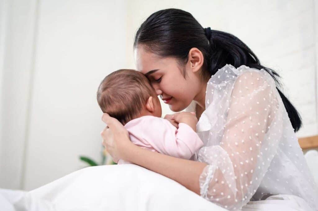 Eine Frau hält ihr Baby sanft im Arm, ihre Stirnen berühren sich zärtlich, während sie gemeinsam auf einem Bett in einem sanft beleuchteten Raum sitzen. Die Umgebung wirkt warm und geborgen und vermittelt Nähe sowie eine zarte Verbundenheit zwischen Mutter und Kind.