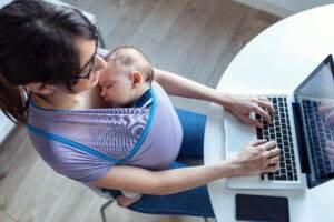 Pretty young mother with her baby in sling working with laptop at home.