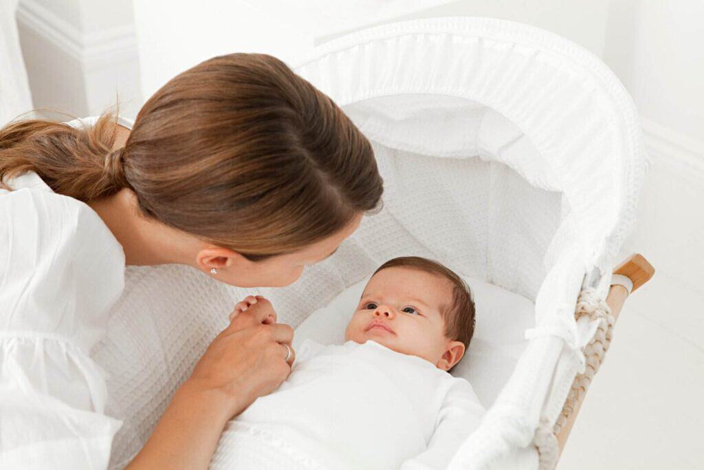 Eine Frau beugt sich sanft über ein Baby, das in einem weißen Stubenwagen liegt, und hält einfühlsam die Hand des Babys. Beide tragen weiße Kleidung. Die helle, ruhige Umgebung vermittelt Geborgenheit und Ruhe.