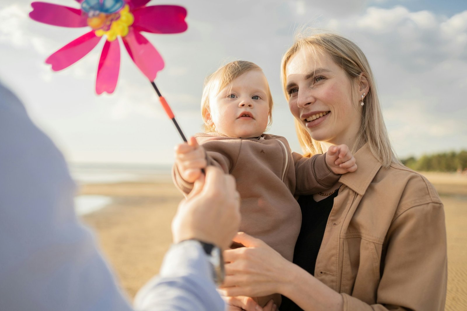 Eine Frau lächelt, während sie ein Baby hält, das nach einem Geburtstrauma-Erlebnis auf ein buntes Windrad blickt, das von einer unsichtbaren Person angeboten wird.