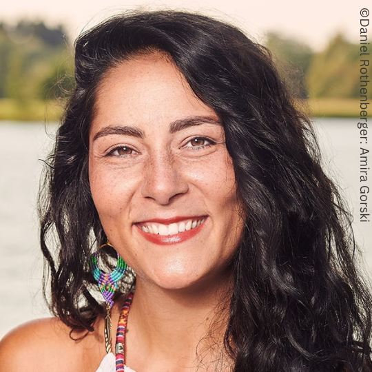 Eine Frau mit langen dunklen Haaren und bunten Ohrringen lächelt in einer Außenkulisse mit Wasser und Bäumen im Hintergrund und erinnert an die heitere Bildsprache, die oft beim TRAUMgeburt nach TRAUMAgeburt-Kongress verwendet wird.