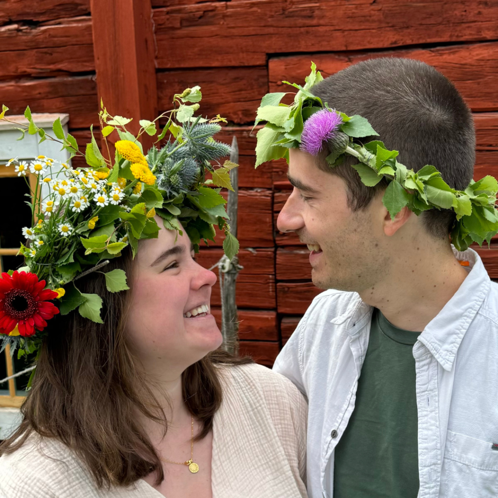 Eine Frau und ein Mann mit Blumenkränzen stehen draußen vor einer roten Holzwand, lächeln und schauen sich an und feiern die Freude über ihre neu gefundene Verbundenheit nach der Teilnahme am TRAUMgeburt nach TRAUMAgeburt-Kongress.