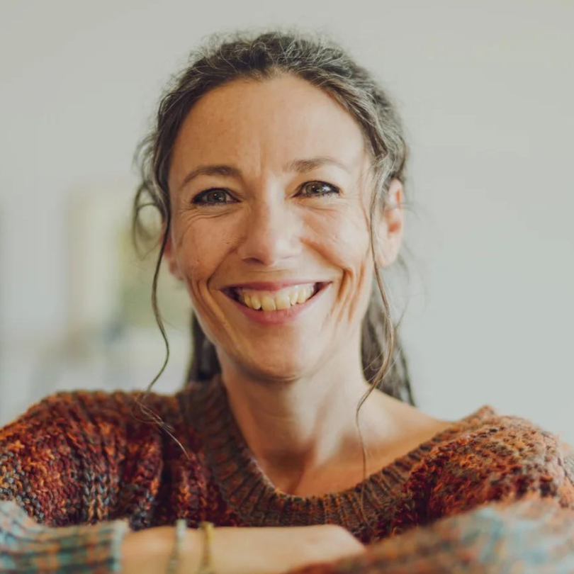 Eine lächelnde Person mit langen Haaren, die einen braunen Strickpullover trägt und die Arme auf einer Oberfläche abstützt, strahlt Ruhe und Wärme aus – ein Beweis für die Stärke der Geburtspsychologie.