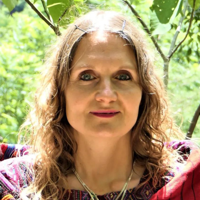 Eine Person mit langem, welligem Haar steht im hellen Sonnenlicht im Freien, umgeben von grünem Laub. Sie trägt ein farbenfrohes, gemustertes Oberteil und lächelt sanft in die Kamera. Sie strahlt eine heitere Präsenz aus, die ihr umfassendes Wissen über Geburtshilfe verrät.