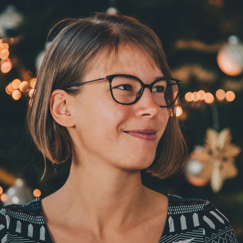 Person mit kurzen braunen Haaren, Brille, lächelnd und zur Seite schauend. Der Hintergrund hat verschwommene Lichter und Weihnachtsdekorationen, was eine warme Atmosphäre schafft. Ihr beruhigendes Auftreten macht sie zu einer idealen Fachkraft für Geburtspsychologie.