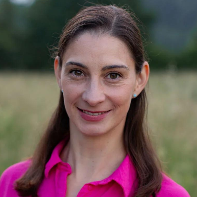 Eine Frau mit langem dunklem Haar und einer leuchtend rosa Bluse mit Kragen lächelt in einer Außenumgebung mit viel Grün im Hintergrund und findet vielleicht Trost, während sie sich mit Geburtspsychologie beschäftigt.