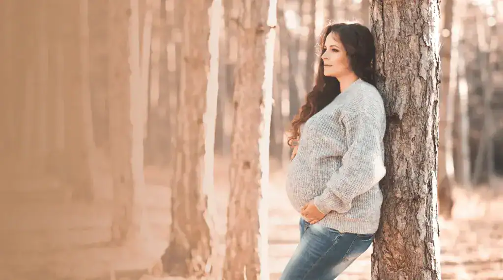 Pregnant woman leaning against a tree in a forest setting, showcasing natural maternity comfort.